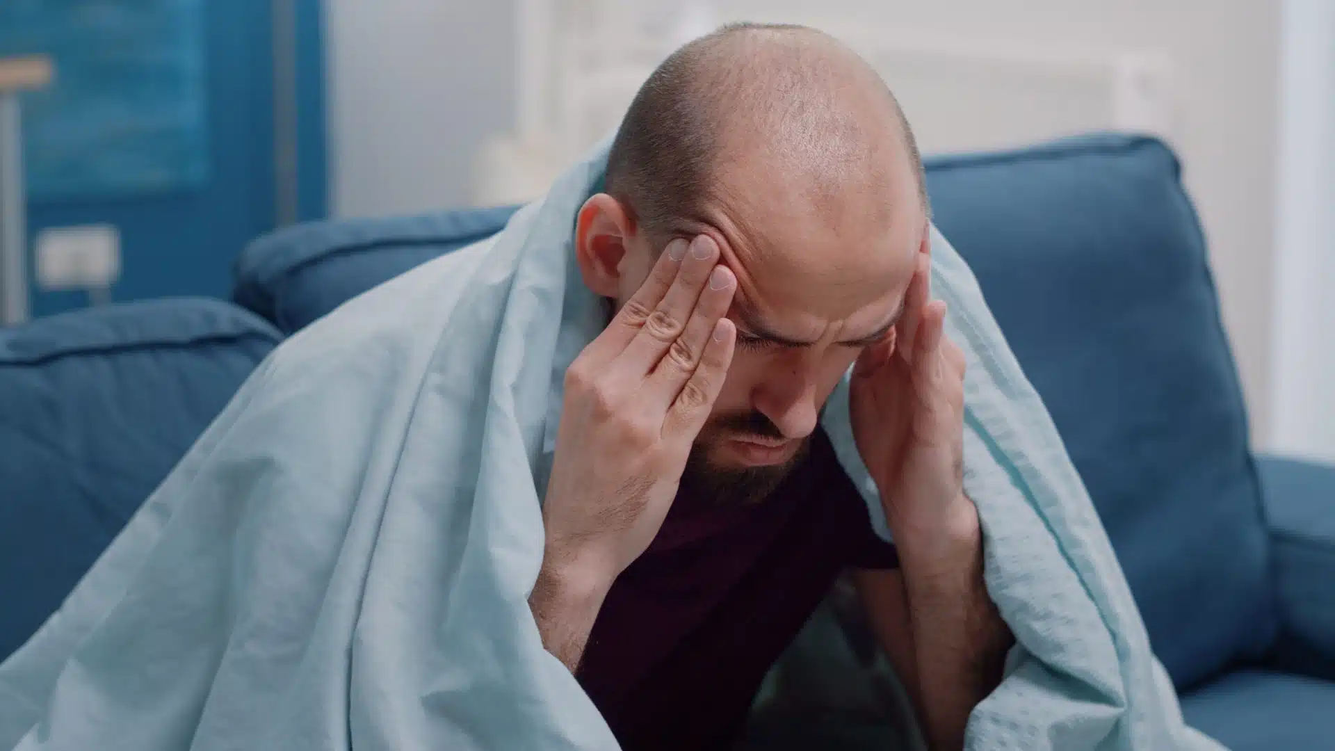Health Hub 21 Man with headache and fatigue sitting on couch covered with blanket pressing temples
