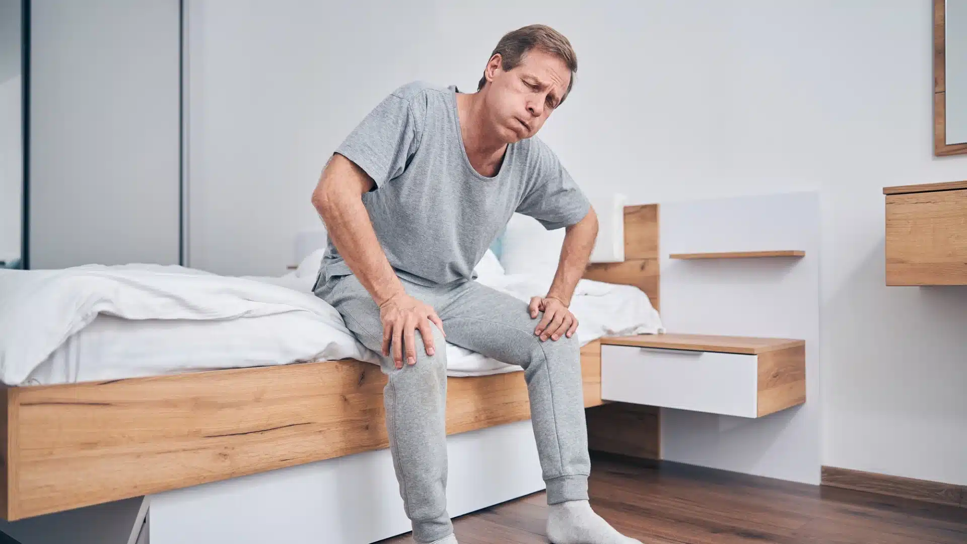Health Hub 11 Middle-aged man sitting on bed holding knees, suffering from joint pain and stiffness in the morning