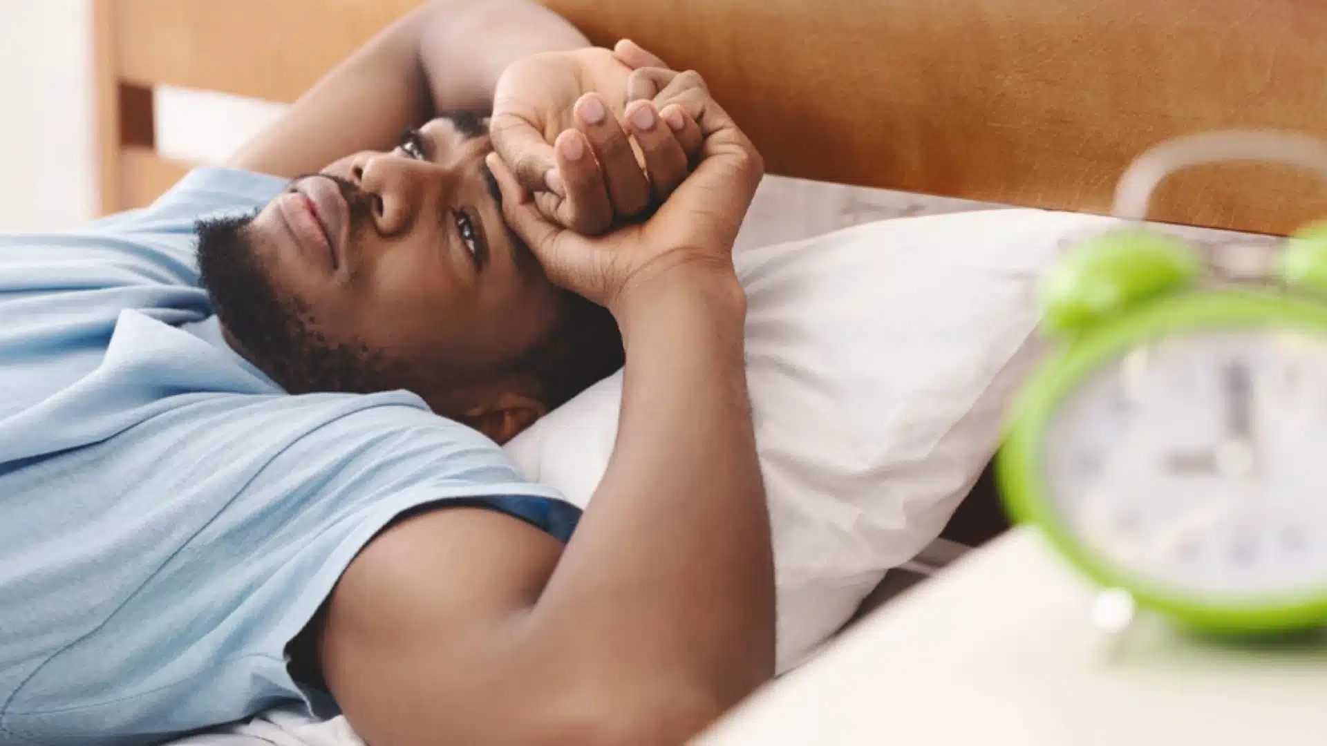 Health Hub 81 Young man lying awake in bed with alarm clock in foreground, experiencing insomnia or sleep disturbance