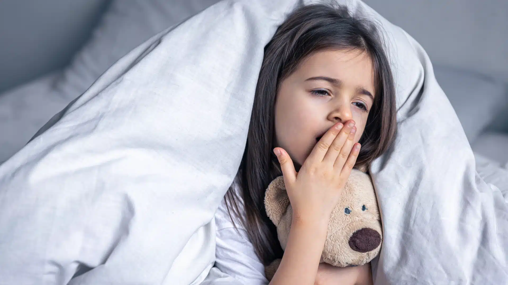 Health Hub 24 Sleepy little girl yawning in bed while holding teddy bear under white blanket