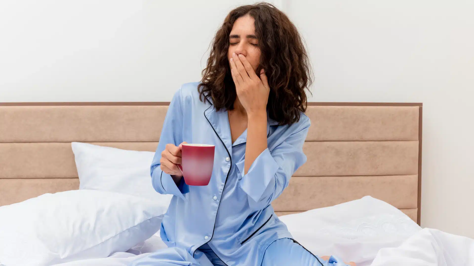 Health Hub 22 Tired woman in blue pajamas yawning and holding coffee cup in bed