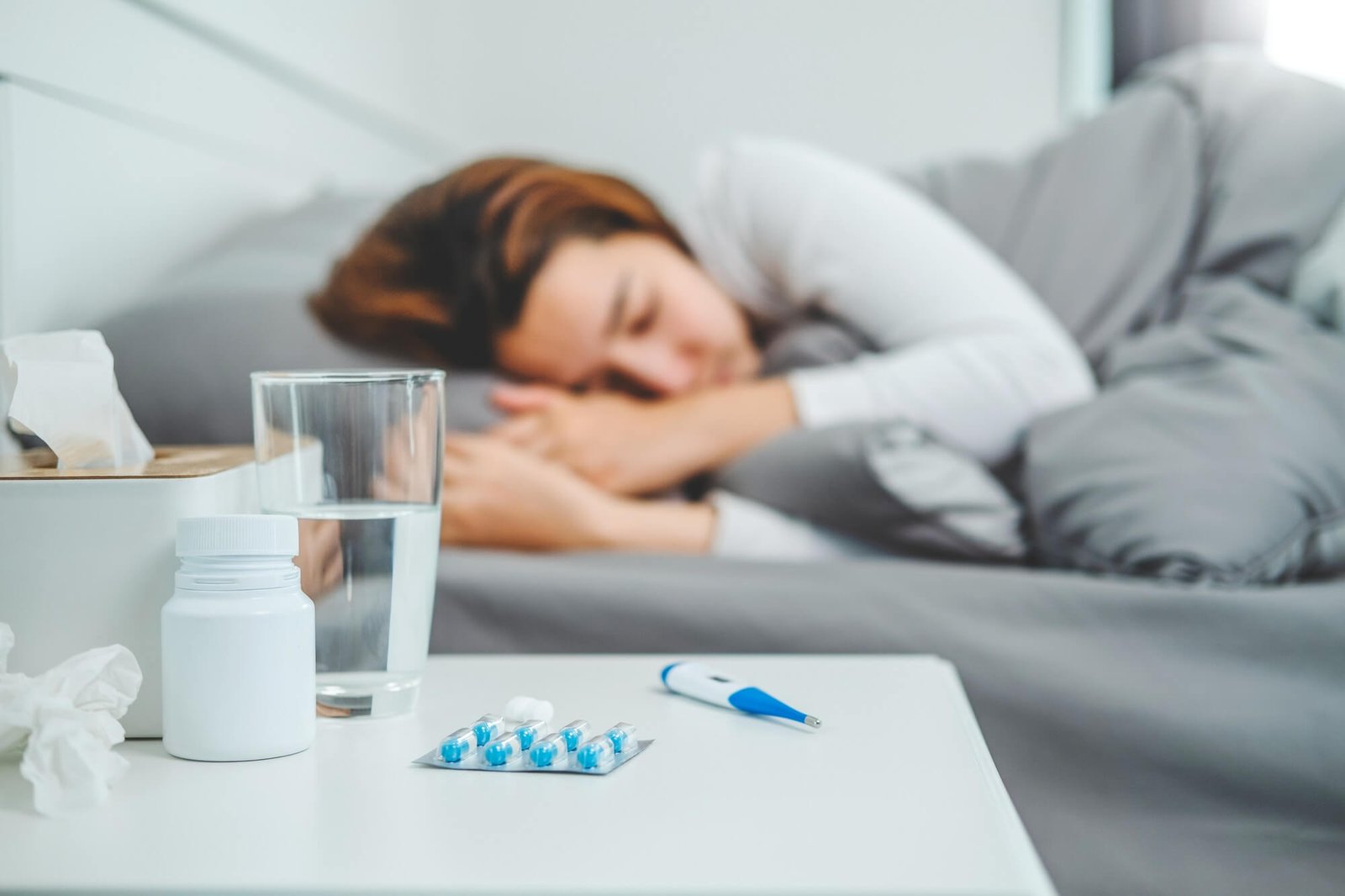 Health Hub 4 Sick woman resting in bed with medicine, glass of water, and thermometer on bedside table