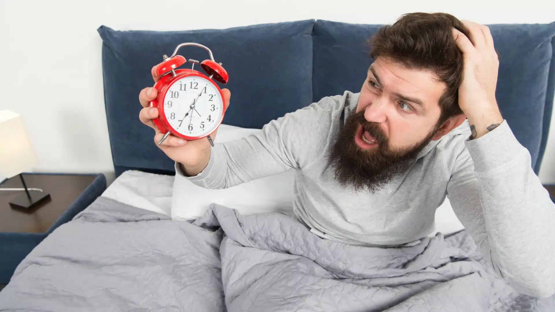 Health Hub 20 Shocked man waking up late in bed holding red alarm clock and looking stressed