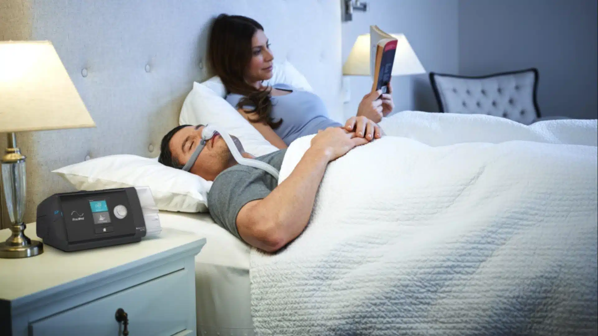 Health Hub 48 Man sleeping with CPAP machine for sleep apnea treatment while partner reads in bed